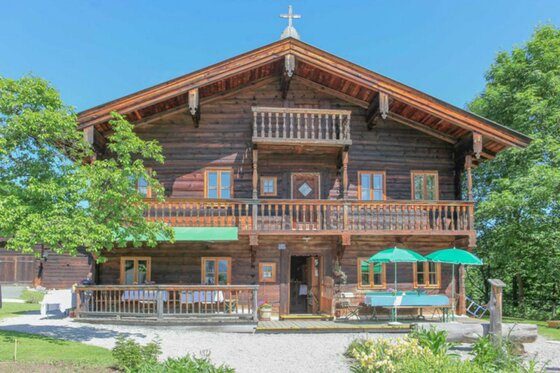 Zweistöckiges traditionelles Holzhaus mit Balkonen, Terrasse und Sonnenschirmen unter blauem Himmel.