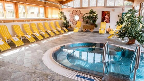 Ein entspannender Wellnessbereich mit einem runden Whirlpool und gelben Liegen. Große Fenster lassen viel Tageslicht herein und schaffen eine ruhige Atmosphäre.
