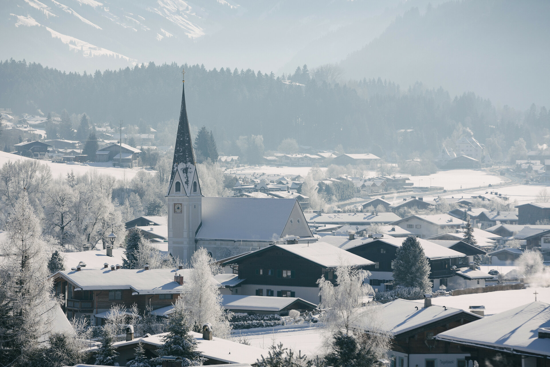 Winterlandschaft in Reith bei Kitzbühel mit Kirche und Häusern, umgeben von verschneiten Bäumen.