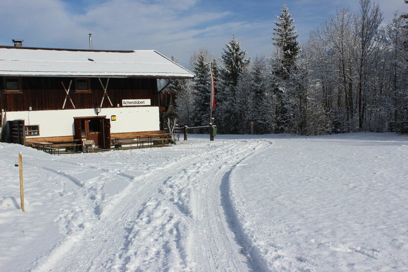 Achenstüberl im Winter Ein verschneites Holzhaus mit weißer Fassade und schneebedecktem Dach neben einer präparierten Winterspur.