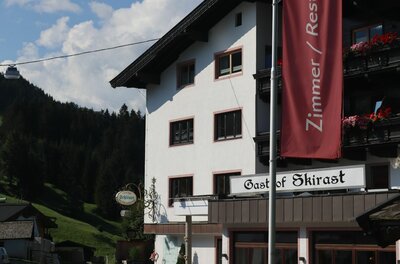 Außenansicht des Gasthof Skirast mit weißer Fassade, Holzbalkonen und roter Fahne vor bewaldetem Berg.