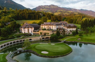 Hotelansicht Schlosshotel Kitzbühel Sommer Ein elegantes Hotel inmitten einer grünen Landschaft und bergigen Kulisse. Ein Golfplatz und ein ruhiger See umgeben das Anwesen.