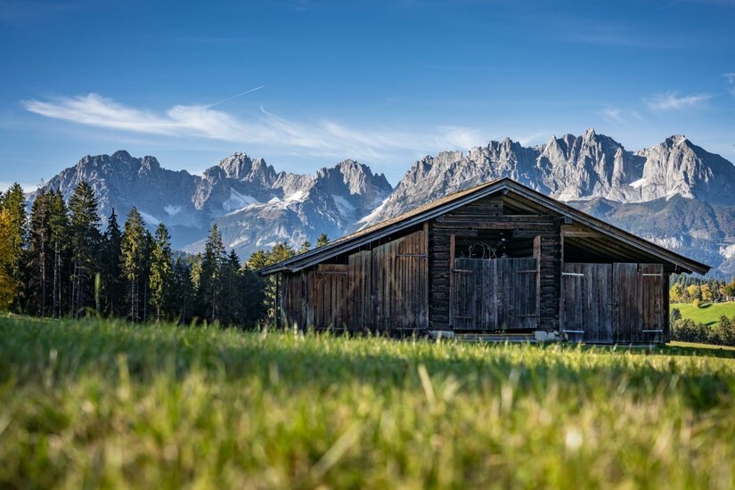 Alte Holzhütte auf einer grünen Wiese vor einem markanten, felsigen Gebirgsmassiv unter blauem Himmel.