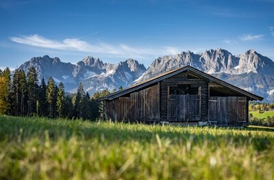Hochzeit - Huestadl <p>Eine rustikale Holzhütte steht vor majestätischen Bergen, dem Wilden Kaiser. Die Landschaft ist grün und einladend mit strahlendem Himmel.</p>