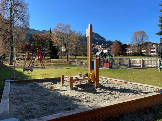 Ein Sandkasten mit Holzumrandung und Spielgeräten auf einer Wiese vor Bergen.
