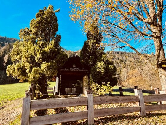Kleine Holzkapelle hinter einem Holzzaun, umgeben von Nadelbäumen und herbstlichem Laub vor bewaldetem Berg.