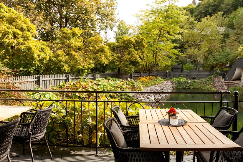 Erika Garten Terrasse Außenterrasse mit Holztischen und schwarzen Korbstühlen vor einem Garten mit Teich und Bäumen.