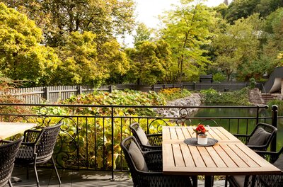 Erika Garten Terrasse Außenterrasse mit Holztischen und schwarzen Korbstühlen vor einem Garten mit Teich und Bäumen.