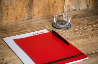 Ein rotes Notizbuch und ein Stift liegen auf einem Holztisch. Neben ihnen steht ein Glas auf einem Untersetzer.