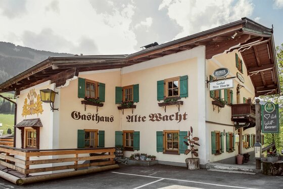 Gasthof Alte Wacht Jochberg Außenansicht des Gasthofs Alte Wacht mit hellgelber Fassade, grünen Fensterläden und Holzbalkon.