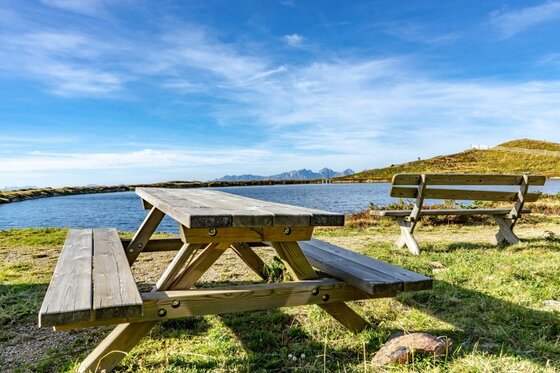 Rastplatz am Pengelstein Holzpicknicktisch und eine Bank an einem Bergsee unter blauem Himmel.