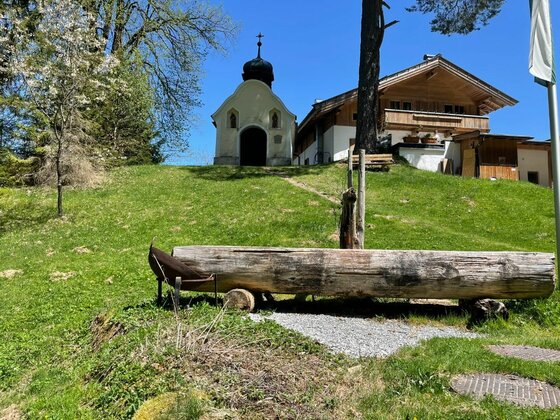 Holzbrunnen auf einer Wiese vor einer kleinen Kapelle und einem Holzhaus unter blauem Himmel.