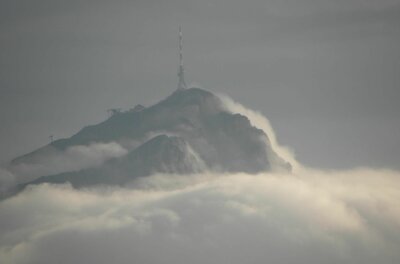 Bergwacht am Gipfel Ein Berggipfel mit Sendemast ragt aus einer dichten Wolkendecke unter grauem Himmel hervor.