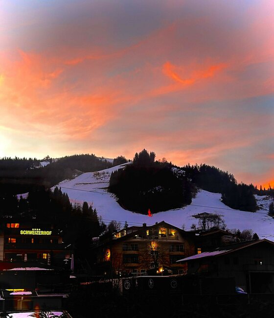 Blick auf beleuchtete Häuser und eine Skipiste vor einem orange-violetten Abendhimmel.