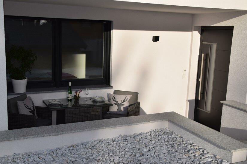 Terrasse Apartment Sabrina Moderne Terrasse mit Sitzgruppe, gedecktem Tisch und Kissen mit Hirschmotiv vor einer weißen Hauswand.