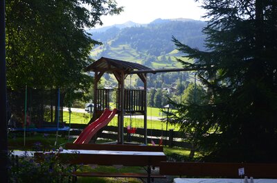 Spielplatz Jodlbühel Holzspielturm mit roter Rutsche und Schaukel vor bewaldeter Bergkulisse.