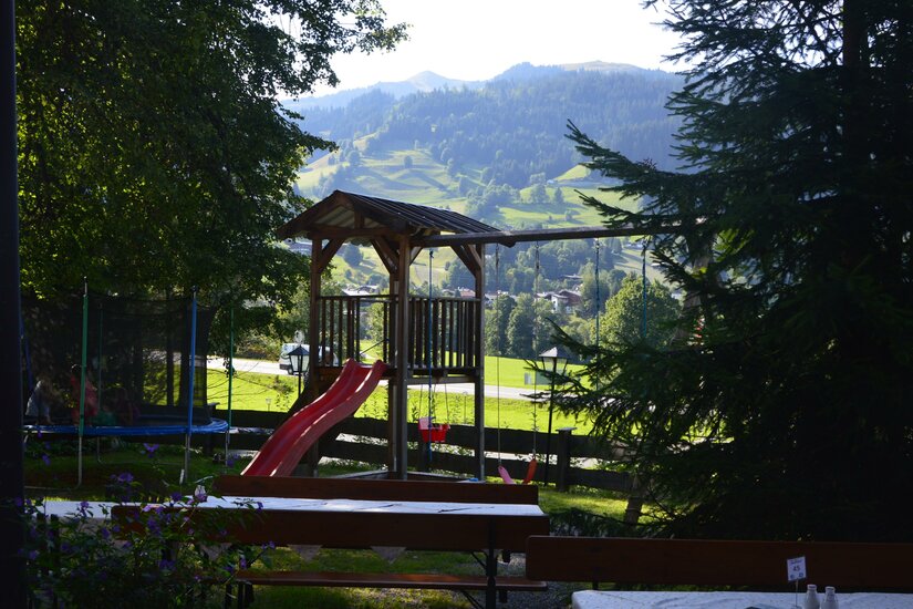 Spielplatz Jodlbühel Holzspielturm mit roter Rutsche und Schaukel vor bewaldeter Bergkulisse.