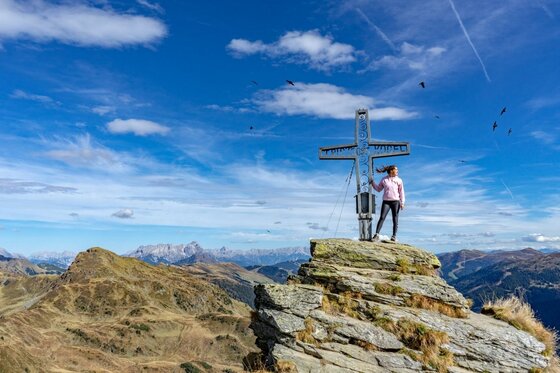 Person steht neben einem metallenen Gipfelkreuz auf einem felsigen Berggipfel vor Alpenpanorama.