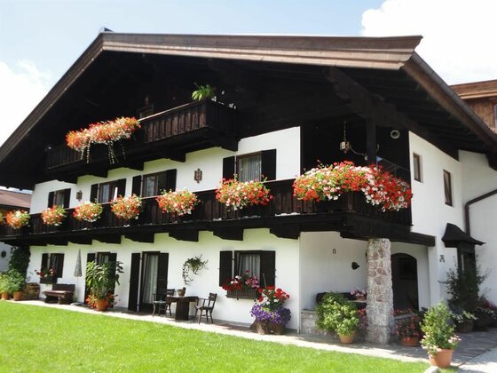 Zweistöckiges weißes Landhaus mit dunklen Holzbalkonen und üppigem Blumenschmuck vor einer Rasenfläche.