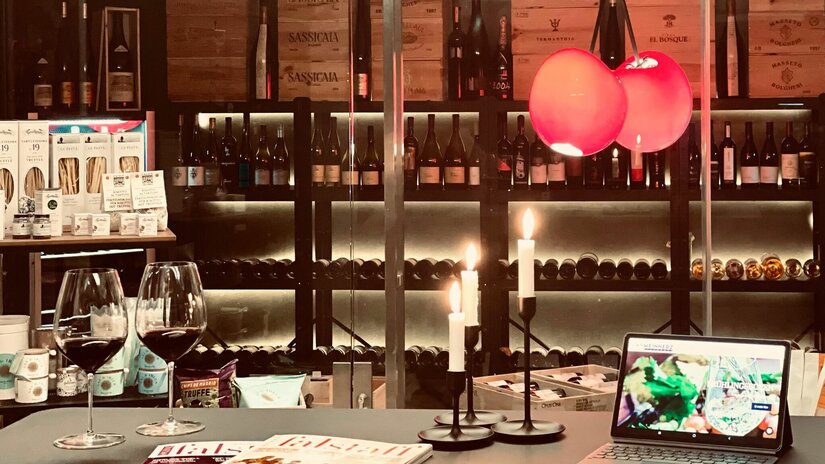 Zwei Gläser Rotwein auf einem Tisch vor einem Weinregal mit Kerzen und einem Tablet.