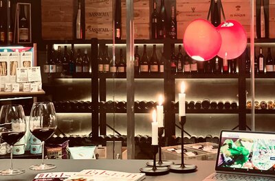 Zwei Gläser Rotwein auf einem Tisch vor einem Weinregal mit Kerzen und einem Tablet.