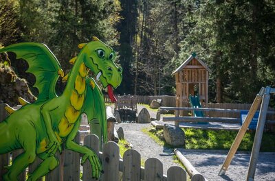 Spielplatz Sintersbacher Wasserfall Grüne Drachenfigur aus Holz an einem Zaun vor einem Waldspielplatz mit Rutsche.