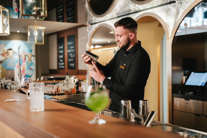 Ein Barkeeper in schwarzer Kleidung bereitet hinter einem Holztresen einen Cocktail mit einem Shaker zu.