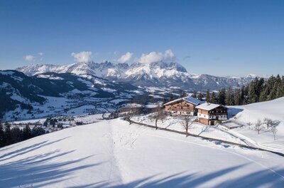 Verschneite Berghütte auf einem sonnigen Hang mit Blick auf ein weites Tal und schneebedeckte Berggipfel.