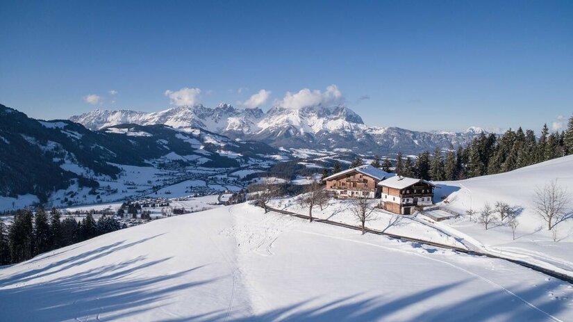 Winterliche Luftaufnahme Hahnenkammblick Verschneite Berghütte auf einem sonnigen Hang mit Blick auf ein weites Tal und schneebedeckte Berggipfel.