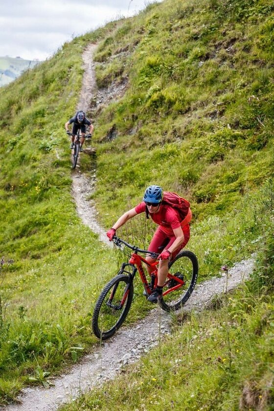 Zwei Mountainbiker fahren hintereinander einen schmalen Schotterpfad an einem steilen Grashang hinunter.
