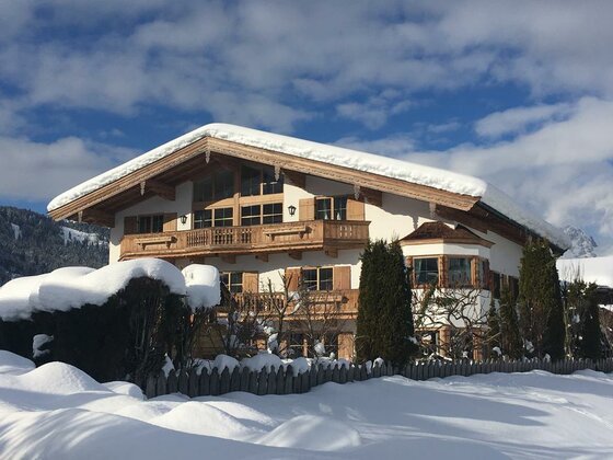 Ansicht Winter Apartment Hahnenkammblick Alpines Wohnhaus mit schneebedecktem Dach und Holzbalkonen vor blauem Himmel.