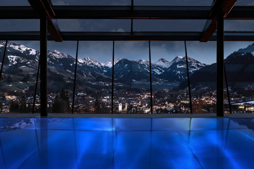 Blick aus einem beleuchteten Innenpool durch eine Glasfront auf ein nächtliches Bergpanorama und eine beleuchtete Stadt.