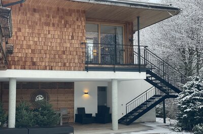 Aufgang zur Streif Lodge Ein modernes Haus mit Holzverkleidung und großen Fenstern. Der Garten ist schneeüberdeckt und im Hintergrund sind schneebedeckte Bäume sichtbar.