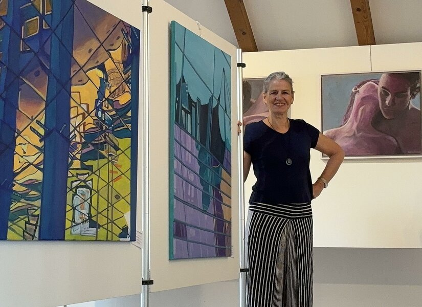 Eine Frau steht lächelnd in einer Kunstgalerie neben mehreren großformatigen, farbenfrohen Gemälden.