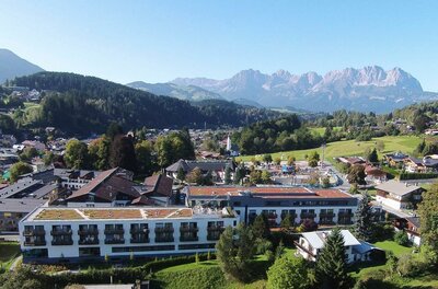 Rehazentrum Kitzbühel Luftaufnahme einer modernen Klinik mit Flachdach vor bewaldeten Hügeln und einem schroffen Gebirgsmassiv.