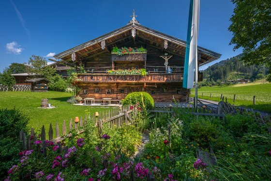 Bauernhausmuseum 2 Ein traditionelles alpenländisches Haus mit Holzbalkon und bunten Blumen im Vordergrund. Die Umgebung ist grün und es herrscht ein klarer blauer Himmel.