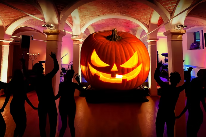 Silhouetten tanzender Personen vor einem großen, beleuchteten Halloween-Kürbis in einem Gewölbekeller.