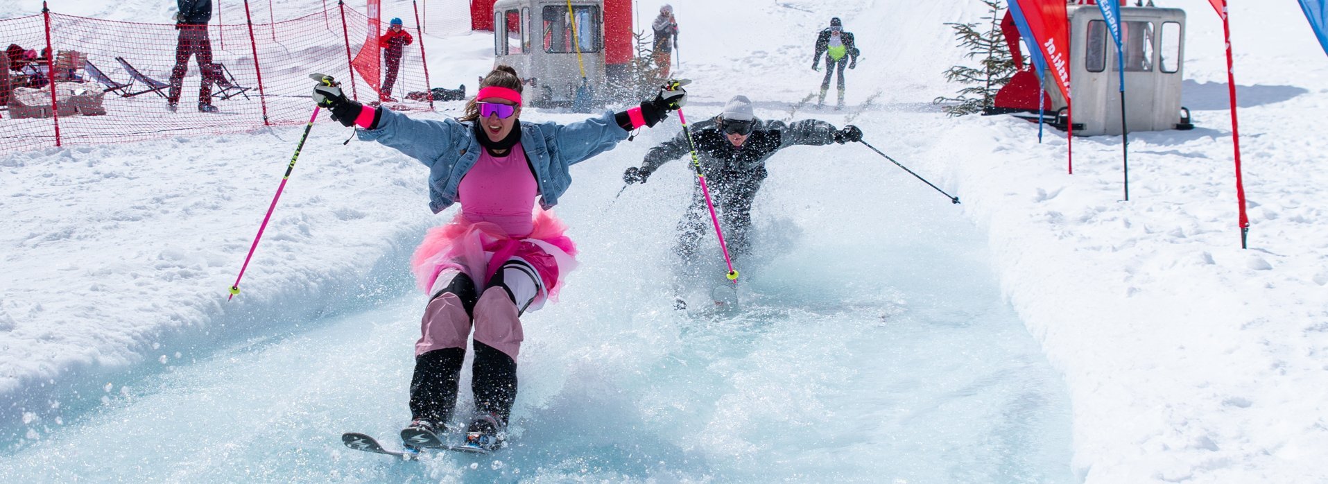 Eine Frau in einem Tütü Rock springt mit Skiern über einem natürlichen Pool auf der Piste 