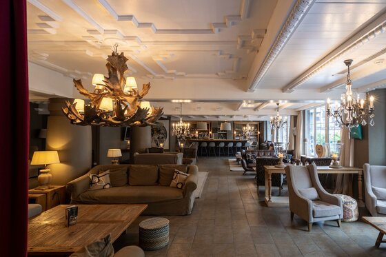 Loungebereich mit Sofas, Sesseln, einem Geweih-Kronleuchter und einer Bar im Hintergrund.