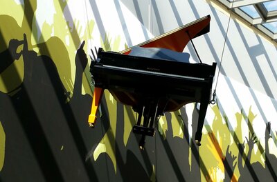 Landesmusikschule Ein schwarzes Klavier hängt an der Wand in einem Raum. Im Hintergrund sind Silhouetten von feiernden Menschen zu sehen.
