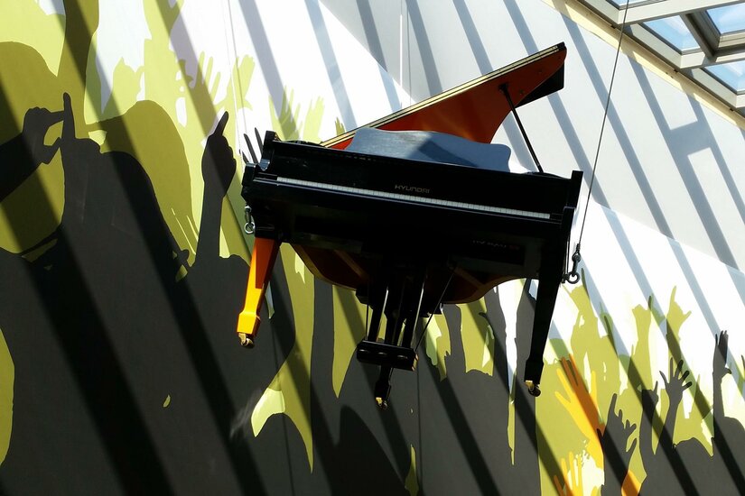 Ein schwarzes Klavier hängt an der Wand in einem Raum. Im Hintergrund sind Silhouetten von feiernden Menschen zu sehen.