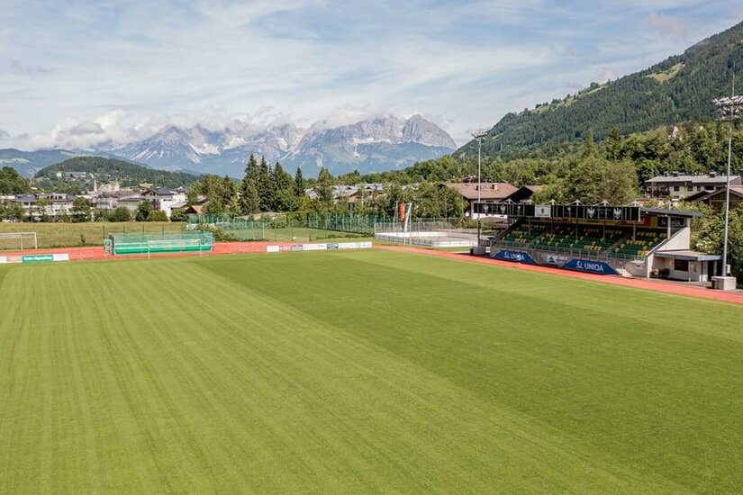 Fußballplatz mit grüner Rasenfläche, kleiner Tribüne und Gebirgskette im Hintergrund unter bewölktem Himmel.