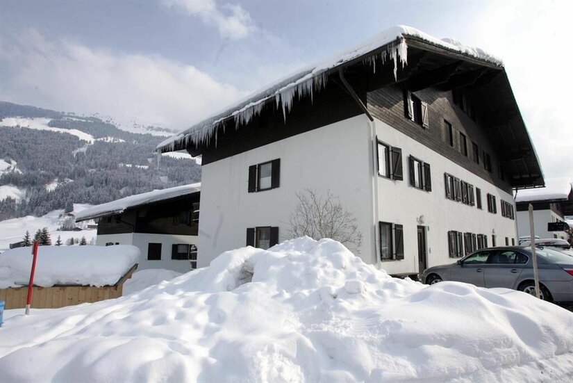 Wohnanlage Wurzhöhe Hausansicht Mehrstöckiges weißes Gebäude mit dunklem Holzdach und Eiszapfen vor verschneiten Bergen.