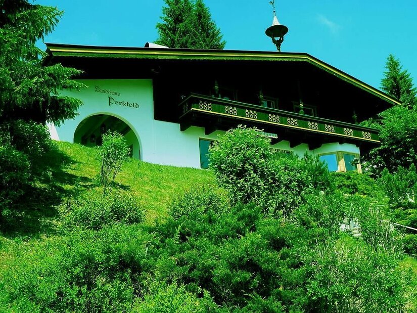 Landhaus Perktold im Sommer Zweistöckiges Landhaus mit dunklem Holzbalkon auf einem grünen Hügel vor Nadelbäumen.