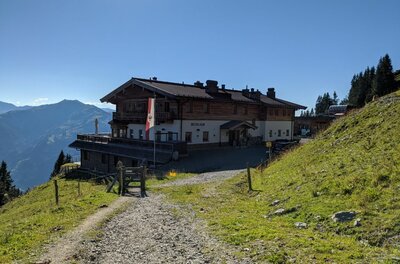 Großes Berggasthaus aus Holz und Stein an einem Schotterweg vor Gebirgspanorama.