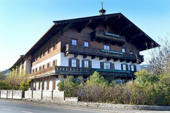 Großes alpines Holzhaus mit weißer Fassade im Erdgeschoss und umlaufenden Holzbalkonen an einer Straße.