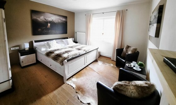 Schlafzimmer mit weißem Doppelbett, Hirschbild an der Wand und zwei schwarzen Sesseln auf Holzboden.