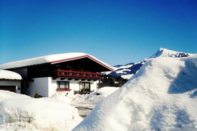 Ein gemütliches Haus in einer verschneiten Landschaft unter klarem blauen Himmel. Hohe Schneehügel umgeben das Gebäude, während Berge im Hintergrund sichtbar sind.