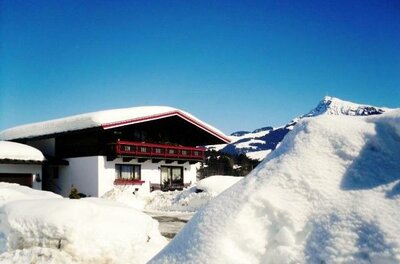 Winter Ein gemütliches Haus in einer verschneiten Landschaft unter klarem blauen Himmel. Hohe Schneehügel umgeben das Gebäude, während Berge im Hintergrund sichtbar sind.