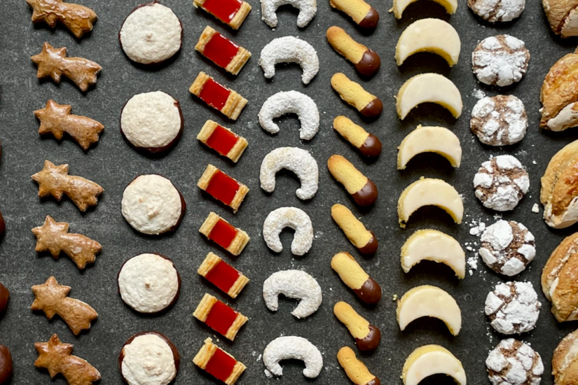 Verschiedene Sorten Weihnachtsplätzchen in Reihen auf dunklem Untergrund angeordnet.
